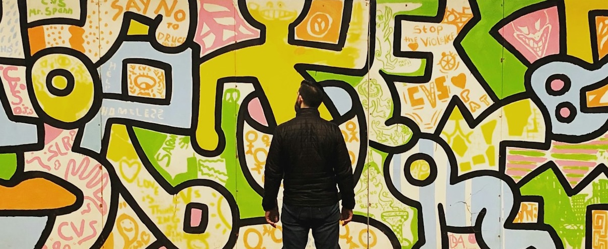 Wall-sized painting (yellow, blue, green, orange abstract people) with man standing in front.
